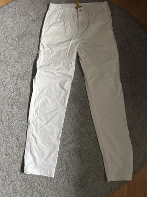 Vita chinos från Polo Ralph Lauren - Säljer ett par klassiska vita chinos från Polo Ralph Lauren. Byxorna har rak passform, elastisk midja och knappar både fram och bak. Tillverkade i mjukt bomullstyg och har diskret logga ovanför ena bakfickan. Perfekta för en clean och stilren look. Storlek M/L