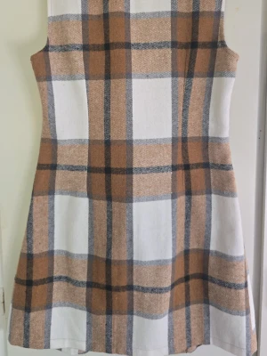 Rutig ärmlös kortklänning med knappar - Snygg ärmlös kortklänning i beige, brun och vit med rutigt mönster. Klänningen har dubbelknäppning framtill, två stora fickor och klassisk krage. Perfekt för dig som gillar retrostil och vill sticka ut.