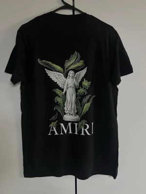 Svart Amiri t-shirt med ängeltryck - Snygg svart t-shirt från Amiri med stort tryck av en ängel och gröna blad på ryggen samt mindre tryck framtill. Klassisk rund hals och korta ärmar. Perfekt för dig som gillar streetwear och vill sticka ut med cool design.