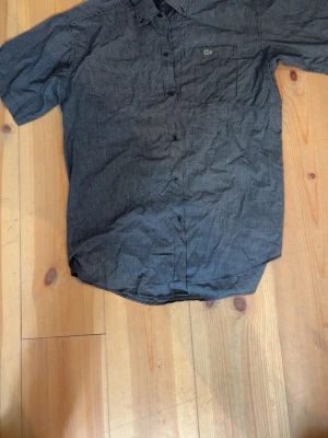 Grå kortärmad skjorta Lacoste - Kortärmad skjorta från Lacoste i grått med diskret rutmönster. Klassisk krage, knappstängning framtill och bröstficka med Lacoste-logga. Perfekt för en avslappnad men stilren look.