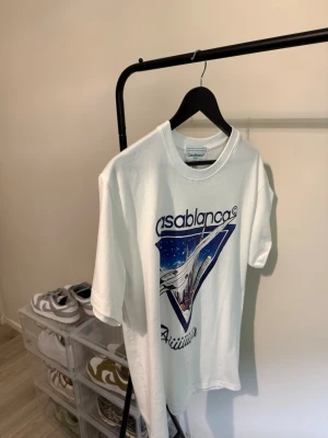 Vit Casablanca t-shirt med tryck - Säljer en vit t-shirt från Casablanca med ett stort grafiskt tryck framtill i blått och lila. T-shirten har rund hals och korta ärmar. Perfekt för dig som gillar streetwear och vill sticka ut med en unik design.