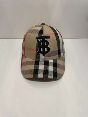 Burberry keps med rutigt mönster - Snygg keps från Burberry i klassiskt beige, svart, vitt och rött rutmönster. Framtill finns en stor svart broderad TB-logga. Kepsen har böjd skärm och justerbar rem baktill. Perfekt accessoar för en trendig streetstyle-look.