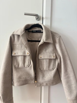 Beige ullkappa från Zara - Säljer en stilren beige kappa från Zara i storlek S. Kappan är tillverkad i ullblandning och har en klassisk krage samt dragkedja framtill. Fodret är ljusbeige och ger en snygg finish. Perfekt för dig som gillar minimalistisk och trendig stil.