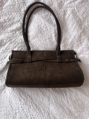 Brun avlång handväska i mockaimitation - Kom gärna med prisförslag!🤗Snygg brun handväska i avlång modell med två långa axelband. Väskan är gjord i mockaimitation och har dekorativa sömmar samt ett stilrent band framtill. Perfekt för dig som gillar klassisk och enkel design.