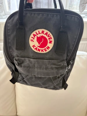 Svart Fjällräven Kånken Mini ryggsäck - Klassisk svart Fjällräven Kånken Mini ryggsäck i slitstarkt material med ikonisk rund röd logotyp framtill. Ryggsäcken har ett stort huvudfack, en mindre ficka med dragkedja framtill och bärhandtag upptill. Perfekt för skolan eller stan.