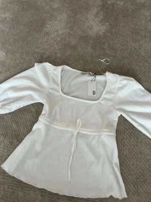 Vit blus med knyt från 157 - Söt vit blus från 157 med fyrkantig ringning och trekvartsärm. Toppen har ett dekorativt knytband i midjan som ger en feminin siluett. Tillverkad i ett lätt och luftigt tyg, perfekt för vår och sommar.