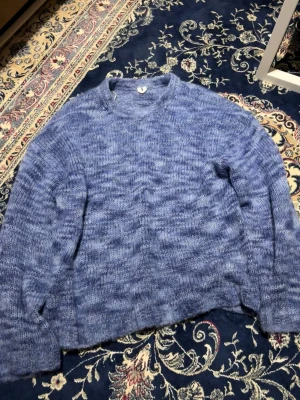 Blå stickad tröja i mohairmix från Arket - Mysig blå stickad tröja med melerad effekt och rund hals. Tröjan har en lös passform och är gjord i en fluffig mohairmix som ger en mjuk känsla. Perfekt för lager på lager och passar till jeans eller kjol.