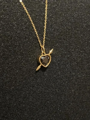 Rebel Heart – Golden Noir - Guldpläterat halsband i rost- och nickelfritt stål. Svart hjärta med pil. Kedja 50 cm, justerbar. Hängets diameter 0,9 cm. 