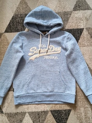 Ljusblå Superdry hoodie - Ljusblå hoodie från Superdry med broderad vit logga på bröstet. Klassisk modell med huva, dragsnören och känguruficka framtill. Mjuk bomullsblandning och ribbade muddar vid ärmslut och nederkant. Perfekt för en avslappnad stil.