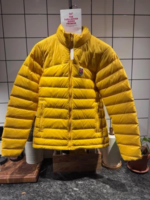 Fjällräven | Expedition pack down jacket W | färg Dandelion -     •    Lätt och mångsidig dunjacka  Inner- och ytterväv av återvunnen nylon  Fylld med 700 fillpower dun, etiskt producerad  Genomsydda dunkanaler  Förstärkt med syntetfyllning, återvunnen polyester, över axlar och i krage  Två handfickor och en innerficka, samtliga med dragkedja  Packas i egen innerficka  Elastiskt band i ärmslut  Dragskojustering i nedre fåll  Frontdragkedja och dragsko designade för att enkelt kunna repareras  Dragkedja i fronten med invändig skyddsklaf 