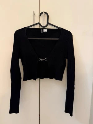 Svart croppad ribbad kofta - Trendig svart croppad kofta med lång ärm och ribbad struktur. Koftan har en djup v-ringning och stängs framtill med en liten silverfärgad hake. Perfekt att styla över ett linne eller t-shirt för en cool look.