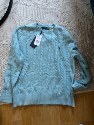 Ljusblå kabelstickad tröja Polo Ralph Lauren - Ljusblå kabelstickad tröja från Polo Ralph Lauren med klassisk rund halsringning och broderad marinblå logga på bröstet. Tröjan har ribbade muddar och nederkant, perfekt för en stilren och avslappnad look.