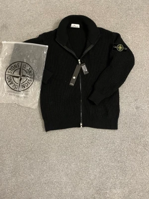 Svart stickad ziptröja Stone Island - Svart stickad ziptröja från Stone Island med ribbstickad struktur och hög krage. Tröjan har dragkedja framtill och den klassiska Stone Island-loggan på vänster ärm. Perfekt för lager-på-lager och streetwear-stil.