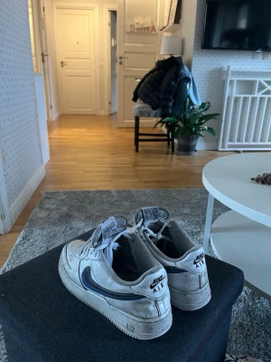 Nike Air Force 1 vita sneakers - Klassiska Nike Air Force 1 sneakers i vitt med grå detaljer och blå Nike-swoosh på sidan. Skorna har lågt skaft, perforerad tå och robust sula. Perfekta för en streetwear-look och passar till det mesta i garderoben.