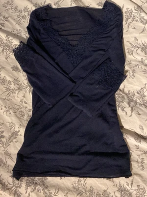 Mörkblå långärmad topp med spets - Snygg mörkblå långärmad topp med v-ringning och spetsdetaljer både vid halsringningen och ärmsluten. Perfekt att styla med jeans eller kjol för en trendig look.