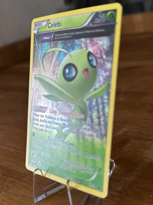 Pokémonkort Celebi holo - Säljer ett holografiskt Pokémonkort med Celebi-motiv. Kortet har gul ram, färgglad bakgrund och tydlig Pokémon-logga på baksidan. Perfekt för samlare eller dig som vill utöka din kortlek med en mytisk Pokémon.