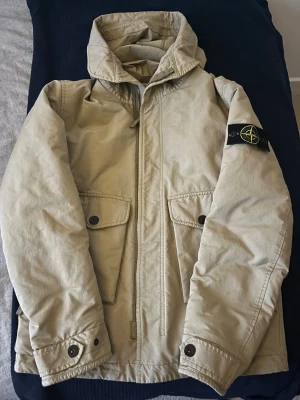 Beige jacka från Stone Island - Snygg beige jacka från Stone Island med hög krage och två stora fickor framtill. Jackan har klassisk logotyp-patch på vänster ärm, knappar vid ärmslut och dragkedja framtill. Perfekt för dig som gillar streetwear och stilren design. Kan gå ner mycket i pris vid snabb affär 