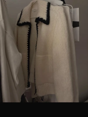 Vit stickad kofta från ZARA - Snygg vit stickad kofta från ZARA med kontrasterande svart kant längs krage och öppning. Koftan har en bred krage och ribbstickade muddar. Perfekt för lager-på-lager och en chill look.