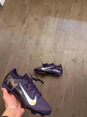 Nike Mercurial KM lila fotbollsskor - Snygga lila Nike Mercurial fotbollsskor med vita och guldiga detaljer samt KM-logga på sidan. Skorna har en lätt och slitstark syntetisk ovandel, samt Air Zoom-sula med rosa dobbar för grymt grepp på planen. Perfekta för dig som vill sticka ut på matchen.