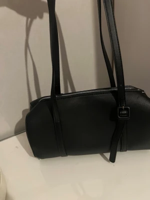 Svart axelremsväska i skinnimitation - Snygg svart handväska i avlång modell med två långa axelband och silverfärgad dragkedja. Väskan har minimalistisk design och detaljer med spänne på framsidan. Perfekt för dig som gillar stilrena accessoarer.