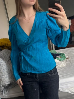 Blå omlottblus med puffärm - Snygg blå omlottblus med puffiga långärmar och knyt i sidan. Djup v-ringning och lätt glansigt material 