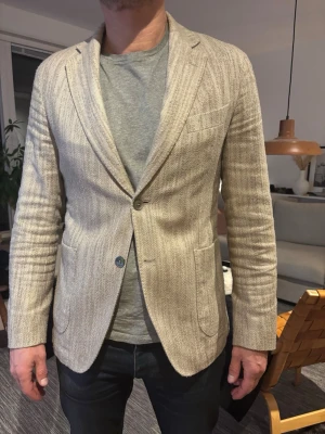 Beige kavaj i linneblandning - Stilren beige kavaj med diskret fiskbensmönster och två knappar framtill. Kavajen har två stora fickor och en bröstficka, samt mjuk passform. Tillverkad i en luftig linneblandning som ger en avslappnad men ändå dressad look. Storlek Slim 50. 