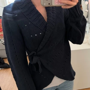 Mörkblå omlottstickad tröja - Säljer en mörkblå stickad omlott-tröja med bred ribbad krage och knytband i sidan. Tröjan har långa ärmar och dekorativa hålstickade detaljer längs ärmar och kropp. Perfekt för dig som gillar en avslappnad men ändå snygg stil.