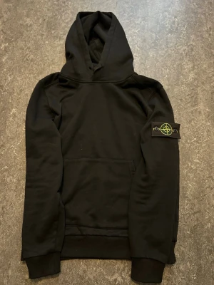 Svart hoodie från Stone Island - Säljer en svart hoodie från Stone Island med klassisk patch på vänster ärm. Hoodien har huva, magficka och ribbade muddar. Tillverkad i mjukt bomullsmaterial och har en clean, stilren look som passar perfekt till streetwear. Storleken på hoodien är S men jag tycker att den motsvarar snarare storleken XS!