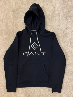 Svart hoodie från GANT  - Svart hoodie från GANT med vit broderad logga och G-symbol på bröstet. Klassisk huva med vita snören och stor magficka framtill. Mjuk bomullskänsla och ribbade muddar vid ärmslut och nederkant. 