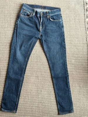 Blå  Nudie Jeans 29,30 - Säljer ett par klassiska blå skinny jeans från Nudie Jeans med fem fickor och orangea sömmar. Jeansen har normal midja och är tillverkade i mjukt denimtyg som ger en snygg passform. Perfekta för dig som gillar en tidlös och clean stil.