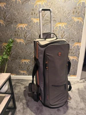 Grå resväska från Timberland - Snygg grå resväska från Timberland med orange detaljer och svarta handtag. Väskan har två hjul, utdragbart teleskophandtag och flera praktiska fack med dragkedja. Tillverkad i slitstarkt tygmaterial och perfekt för längre resor.