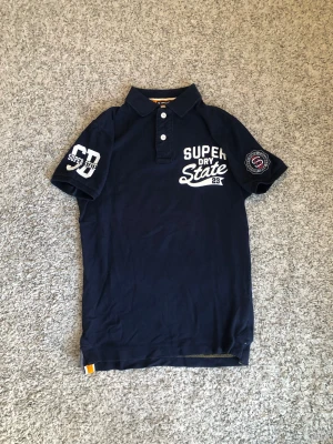 Mörkblå piké från Superdry - Snygg mörkblå pikétröja från Superdry med vita tryckdetaljer på bröstet och ärmarna. Klassisk krage med tre knappar framtill och korta ärmar. Perfekt för en avslappnad och sportig stil.