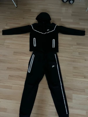  Nike tech M - Svarta mjukisbyxor från Nike med vita detaljer längs sidorna och vid fickorna. Byxorna har resår och dragsko i midjan samt Nike-logga på låret. Matchar perfekt med en hoodie för en sportig och avslappnad stil.