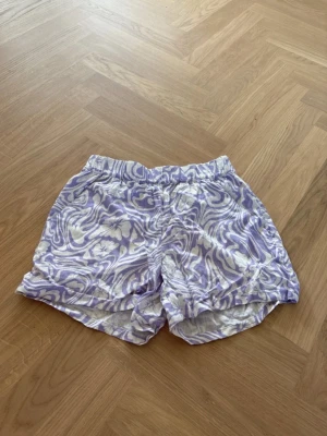 Lila blommiga shorts med resår - Säljer ett par lätta shorts med lila och vitt blommönster och swirliga linjer. Shortsen har resår i midjan och är gjorda i ett tunt, luftigt material som passar perfekt för sommaren.