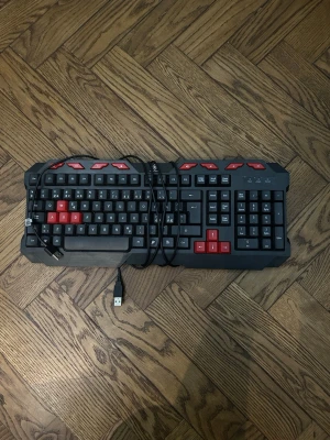 Svart gamingtangentbord med röda detaljer - Säljer ett svart gamingtangentbord med röda knappar på WASD och piltangenterna. Tangentbordet har USB-anslutning och en robust design med extra röda knappar upptill. Perfekt för dig som vill ha en cool look till din setup.