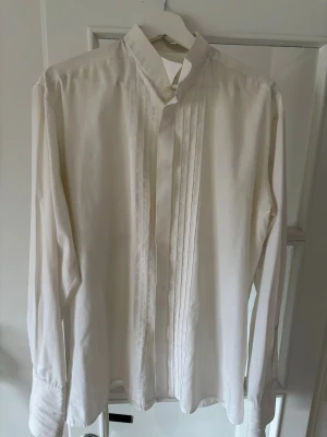 Vit långärmad smokingskjorta med ståkrage - Klassisk vit skjorta med ståkrage och veckade detaljer framtill. Skjortan har långa ärmar och dolda knappar för en clean look. Perfekt till en stilren smoking