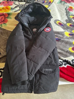 Svart dunjacka från Canada Goose - Säljer en svart dunjacka från Canada Goose med klassisk patch på bröstet och dragkedja framtill. Jackan har huva, två stora fickor med lock och diskret logga på ena fickan. Perfekt för kalla vinterdagar och riktigt varm.