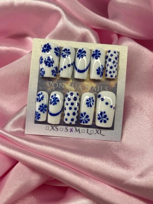 Bleu (M);Medium Square;Curved - Snygga handgjorda press-on naglar i vit bas med handmålade blå blommor och prickar. Naglarna har en medium,  fyrkantig form och är perfekta för dig som vill ha en unik och trendig look. I storlek M: 16 mm, 12 mm, 12,5 mm, 11,5 mm, 10 mm och designen är handmålad och riktigt detaljerad.