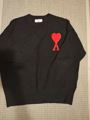 Svart stickad tröja från Ami Paris - Svart stickad tröja från Ami Paris med röd hjärtlogga och bokstaven A på bröstet. Tröjan har rund halsringning, ribbstickade muddar och nederkant. Perfekt för dig som gillar stilrena plagg med en twist.