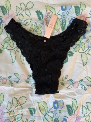 Svarta spets-trosor från Victoria's Secret - Aldrig använda eller testade, köpte fel storlek. Snygga svarta trosor i brasiliansk modell från Victoria's Secret. Trosorna är gjorda i mjuk spets med blommigt mönster och har en liten rosa detalj framtill. Perfekta för dig som gillar både komfort och stil. ☺️
