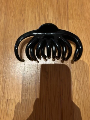 Svart hårklämma i plast - En klassisk svart hårklämma i glansig plast med böjda, breda klor som håller håret på plats. Perfekt för att snabbt sätta upp håret i en snygg och enkel frisyr. Stilren och tidlös design som passar till många looks.