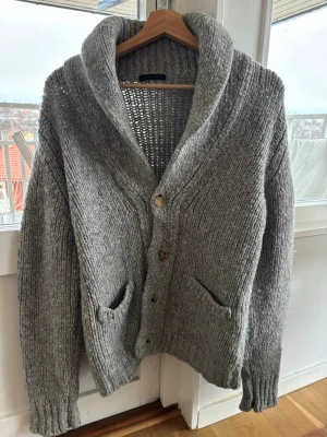 Grå stickad kofta från Roberto Collina - Mysig grå stickad kofta från Roberto Collina med stora knappar, två fickor framtill och bred sjalkrage. Koftan har en chunky känsla, men riktigt skön. Storlek 48. Hör av er om ni har frågor. 