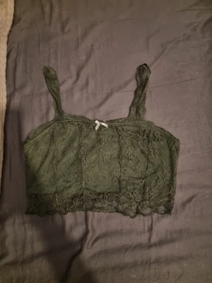 Grön spets crop top med rosett - Söt grön linnetopp i spets med smala axelband och liten rosett framtill. Toppen har en croppad passform och fina spetsdetaljer längs nederkanten. Perfekt att styla med höga jeans eller kjol. Inte helt hundra på storlek men skulle gissa på M.