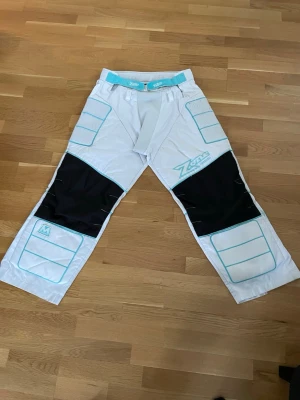 Zone målvaktsbyxor vit/blå - Säljer ett par vita målvaktsbyxor från Zone med ljusblå detaljer och svarta förstärkningar på knäna. Byxorna har vadderade partier för extra skydd och dragkedja vid benslut. Perfekta för innebandy och ger bra rörelsefrihet. Finns hål vid båda dragkedjorna men hindrar inte funktionen på byxan