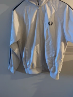 Vit Fred Perry ziptröja - Snygg vit ziptröja Fred Perry med svart logga på bröstet och svarta ränder längs axlarna. Tröjan har hög krage, dragkedja framtill och ribbade muddar. Perfekt för en sportig och stilren look.
