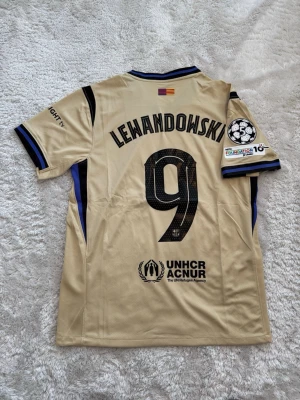 Barcelona Lewandowski matchtröja guld - Snygg Barcelona matchtröja i guld med Lewandowski och nummer 9 på ryggen. Tröjan har svarta detaljer, Champions League-märke på ärmen och Spotify-logga på bröstet. Tillverkad i lätt och ventilerande material, perfekt för fotbollsträning eller match.