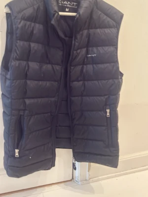 Mörkblå dunväst från GANT - Snygg mörkblå dunväst från GANT med quiltad design och två dragkedjeförsedda fickor framtill. Västen har hög krage och diskret GANT-logga på bröstet. Perfekt att slänga över en hoodie eller tröja för extra värme. Materialet är lätt och smidigt. Ny pris är 2000kr+