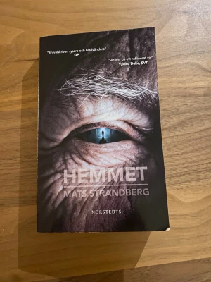 Hemmet av Mats Strandberg - Skrämmande och gripande roman av Mats Strandberg, utgiven av Norstedts. Omslaget är mörkt med ett närbild på ett öga och en siluett i pupillen. Boken är en pocket och handlar om kärlek, vänskap och skräck på ett äldreboende.