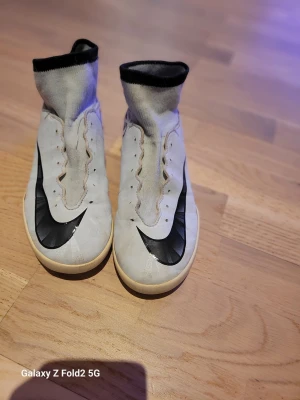 Nike CR7 fotbollsskor - Säljer ett par vita Nike Mercurial fotbollsskor med CR7-tryck på hälen och stora svarta swoosh-loggor på ovansidan. Skorna har högt skaft i stickat material och svart kant upptill. Perfekta för dig som vill sticka ut på planen.