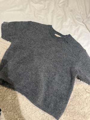 Grå stickad kortärmad tröja - Mysig grå stickad tröja med korta ärmar och rund hals. Tröjan har en avslappnad passform och är tillverkad i ett mjukt, luddigt material. Perfekt att styla lager på lager eller bära som den är.